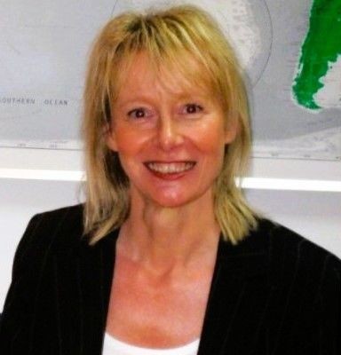 Smiling person with short blond hair wearing a black jacket, in front of a map.